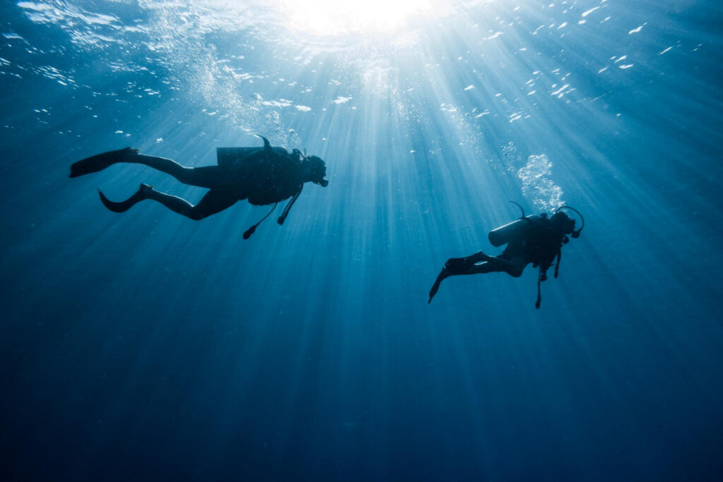 Dois mergulhadores nadando nas águas cristalinas do oceano com raios de sol atravessando a água no Alentejo.