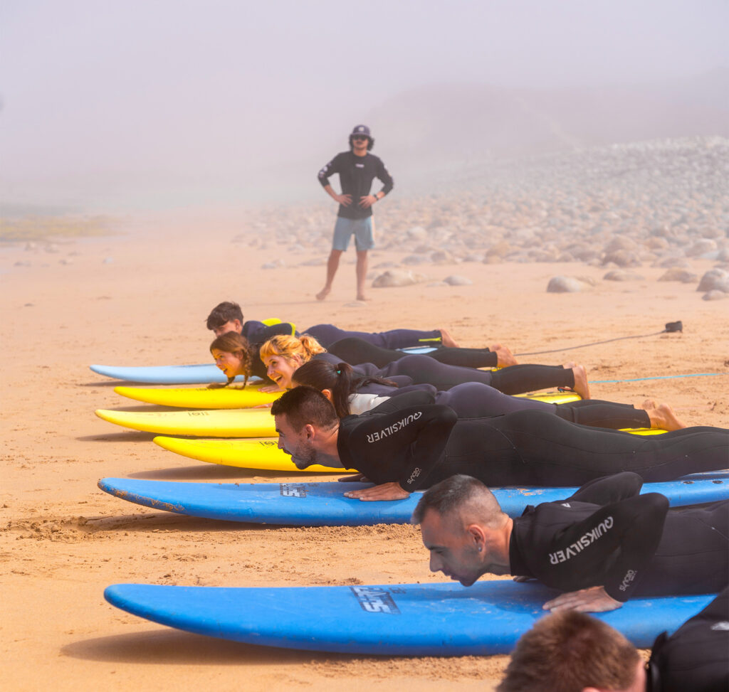 Students in wetsuits practicing the take-off maneuver on the beach during a group lesson with an instructor in the background in Porto Covo.