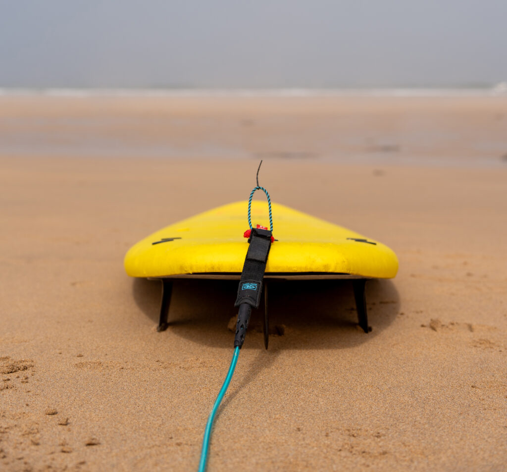 The back of a yellow surfboard with a blue leash and fins, resting on the sand on the beach facing the ocean in Porto Covo.