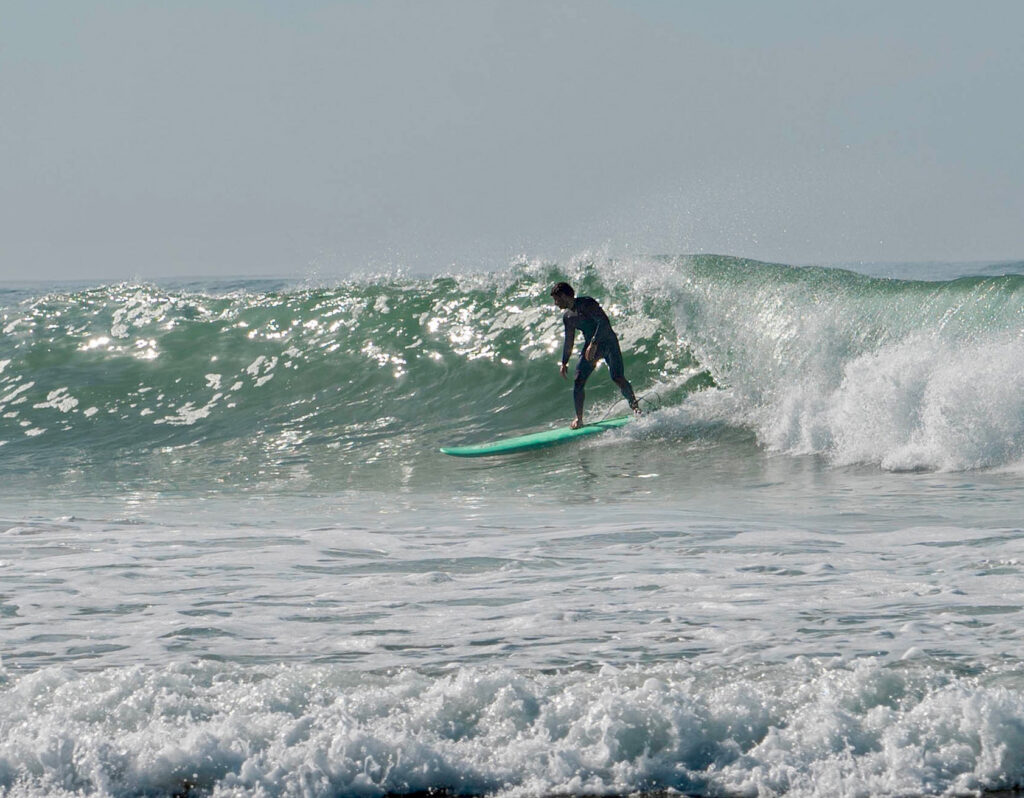 A surfer in a dark wetsuit gliding on a green, crystal-clear wave with a longboard on a beach in Porto Covo.